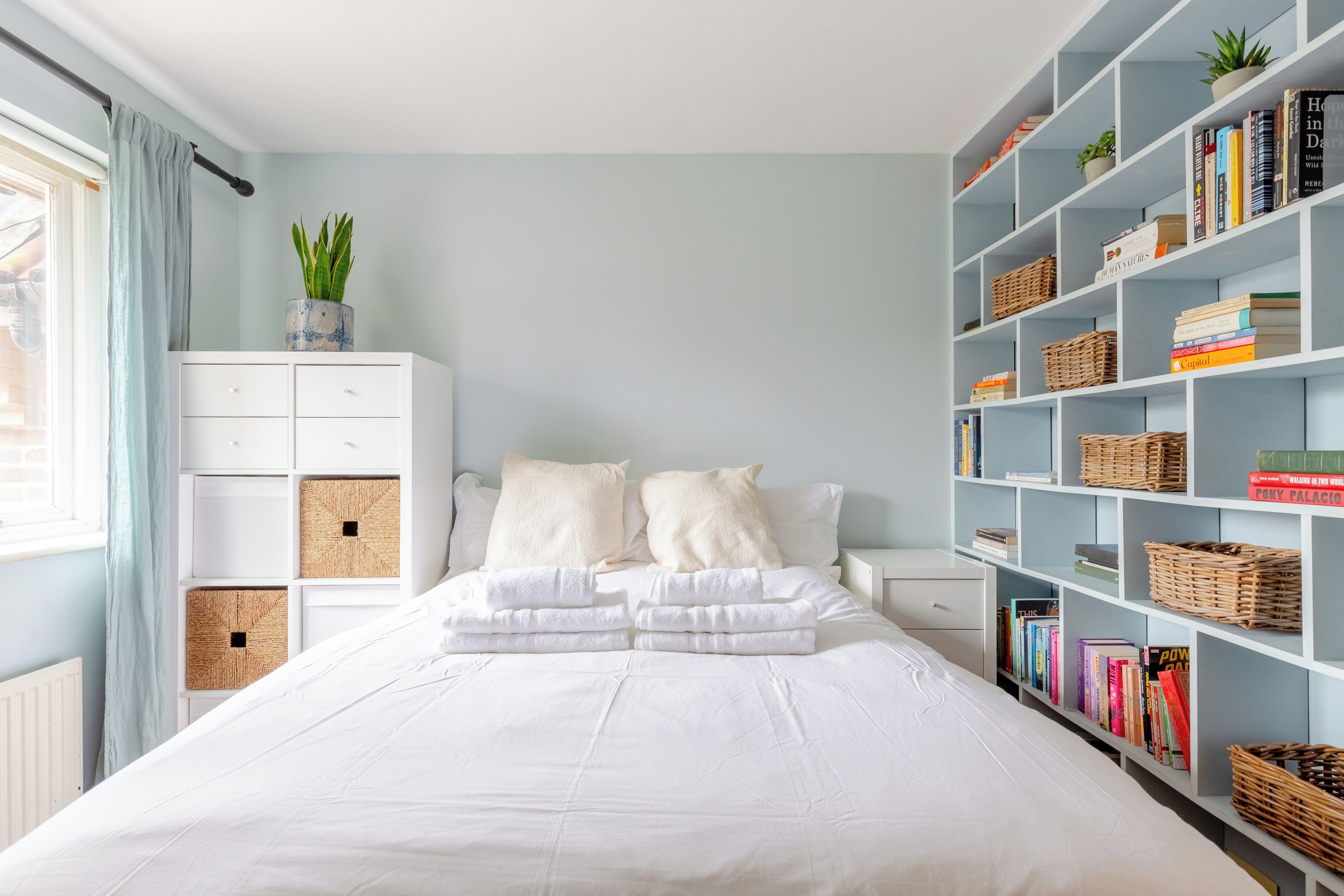 Una camera da letto con un letto, biancheria da letto bianca, un comodino, una libreria e una finestra con tende.