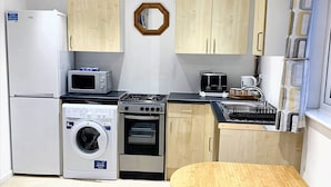 A compact kitchen with a refrigerator, microwave, washing machine, oven, and a small dining table. - Lovely one bedroom flat (Wooburn Green, Bourne End)