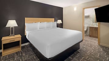 Une chambre d’hôtel avec un grand lit, deux tables de chevet avec des lampes, une petite table en bois, un téléviseur fixé au mur et une salle de bain visible à travers une porte ouverte.