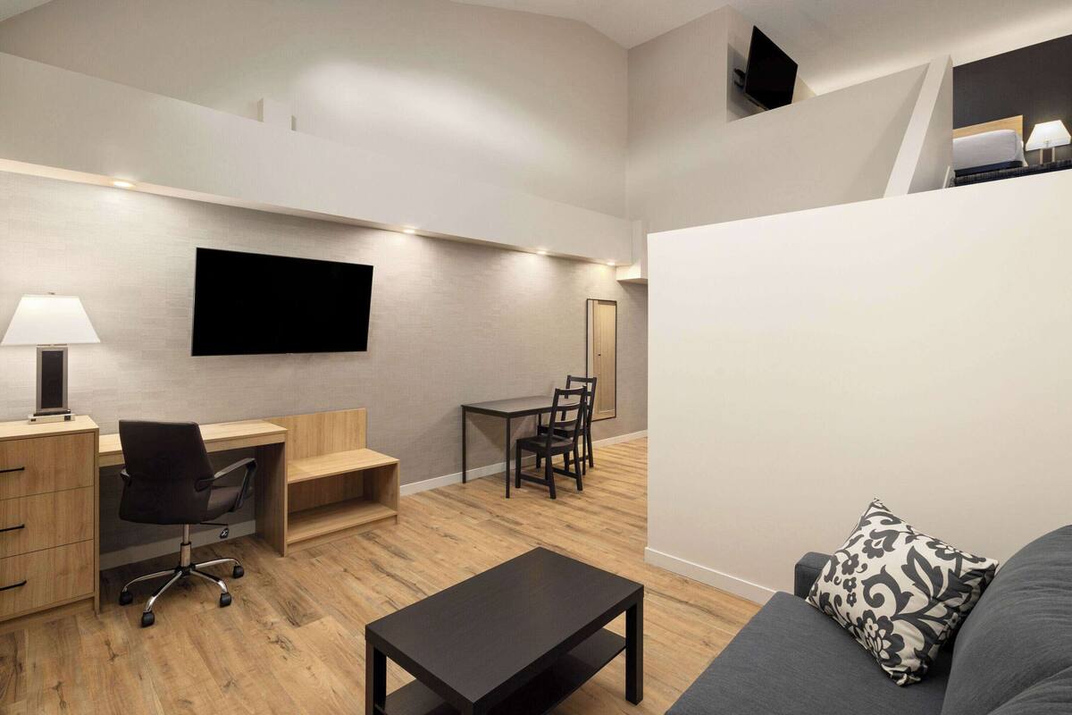 Een moderne woonkamer met een flatscreen televisie aan de muur, een houten bureau met stoel, een bank met een gekleurd kussen en een kleine tafel.