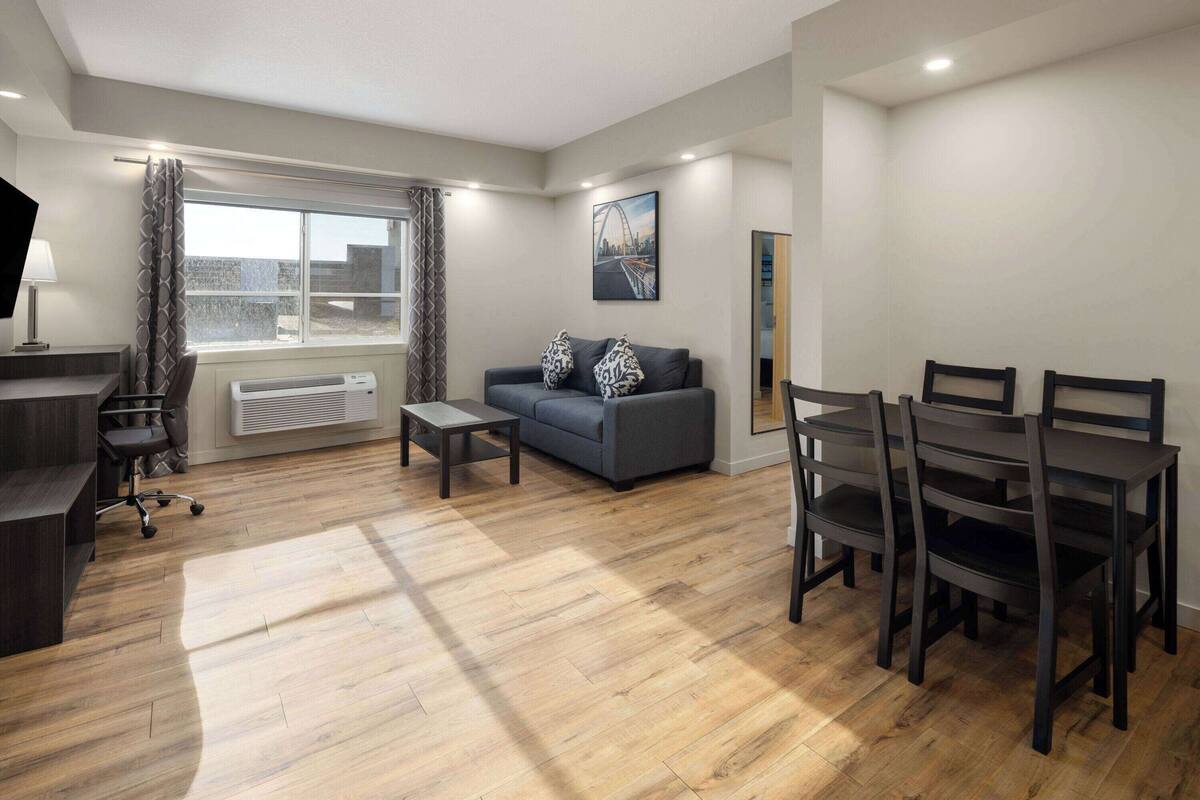 Een moderne woonkamer met een grijze bank, decoratieve kussens, een salontafel, een eettafel met stoelen, een televisie en een groot raam met gordijnen.