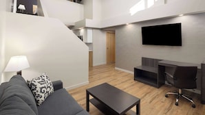Una sala de estar moderna con un sofá gris, una mesa de centro, un televisor montado en la pared y una escalera que lleva a un nivel superior.
