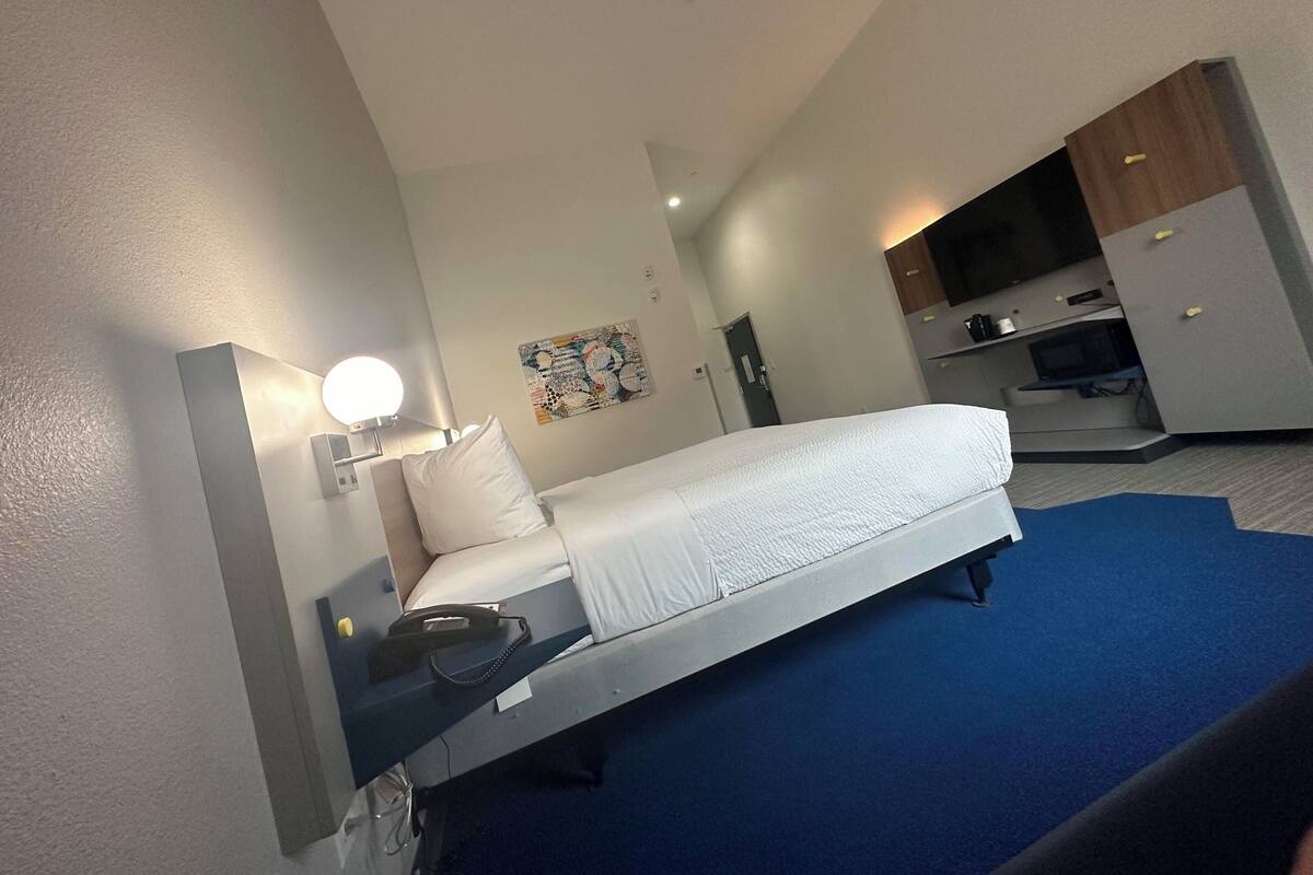 A hotel room with a bed, bedside table, lamp, and a small desk with a chair.