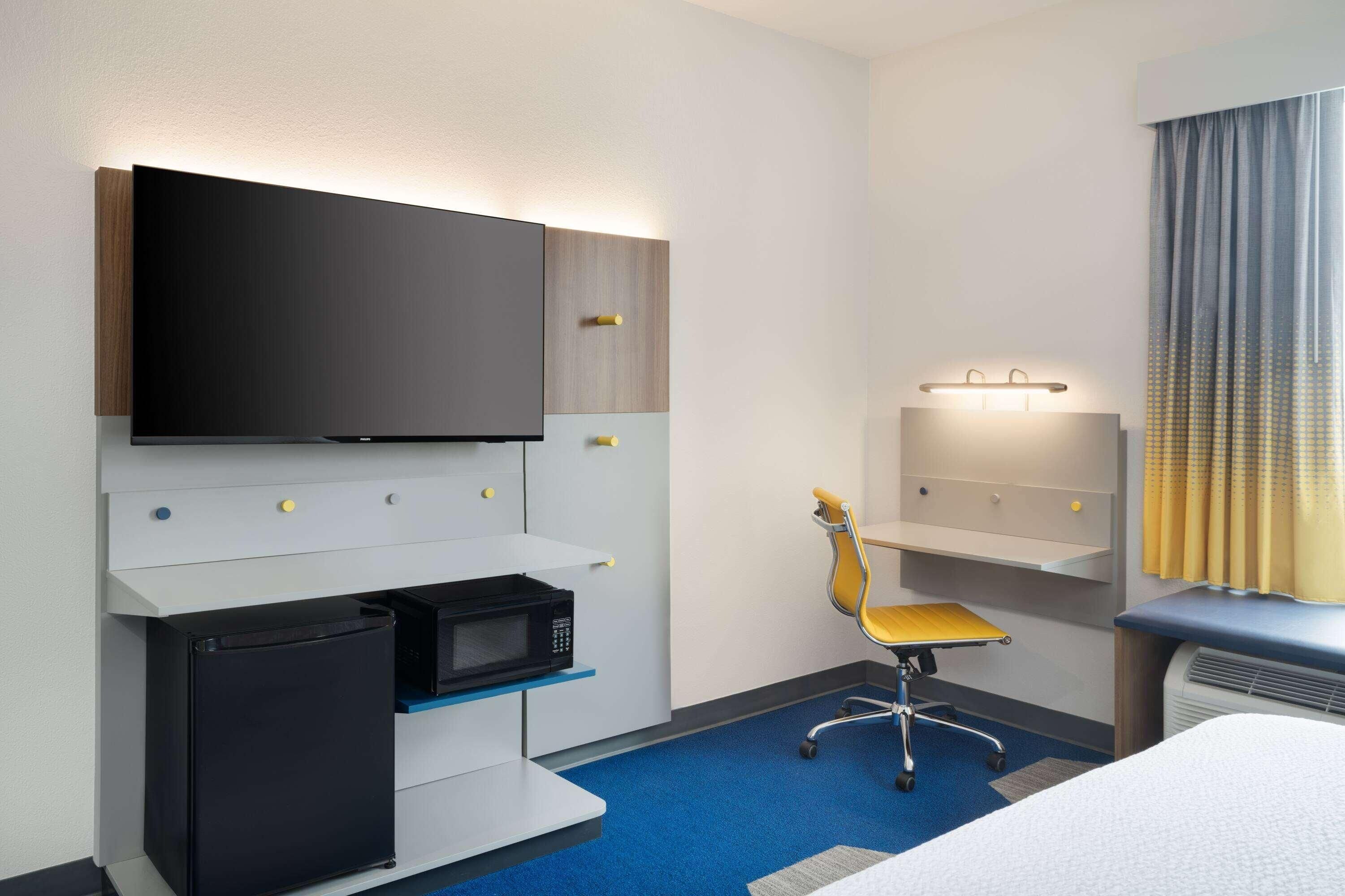 A hotel room with a flat-screen TV, a microwave, a small refrigerator, a desk with a chair, and a window with curtains.