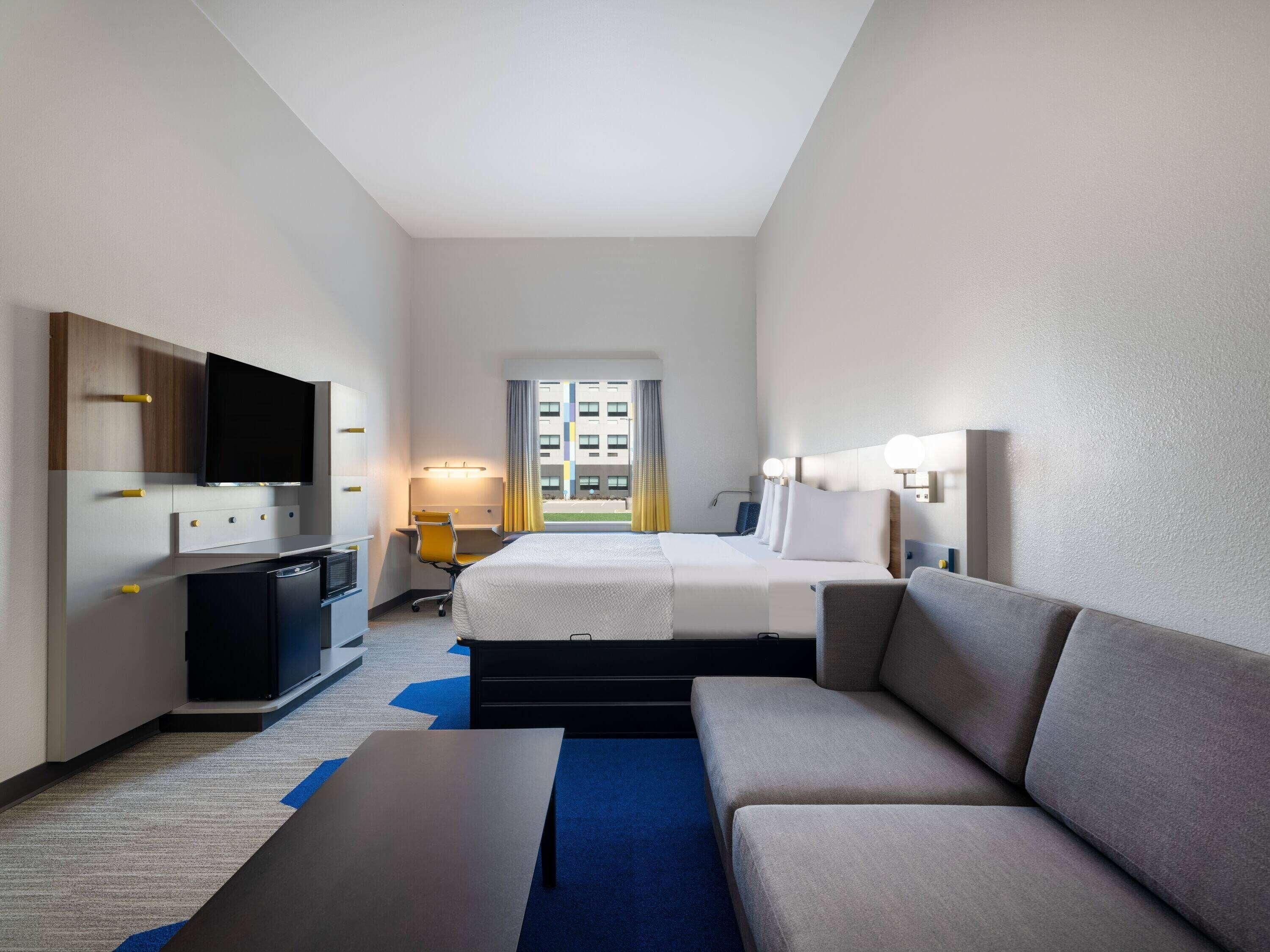 A hotel room with a bed, a sofa, a desk, a TV, and a window with a view of buildings.
