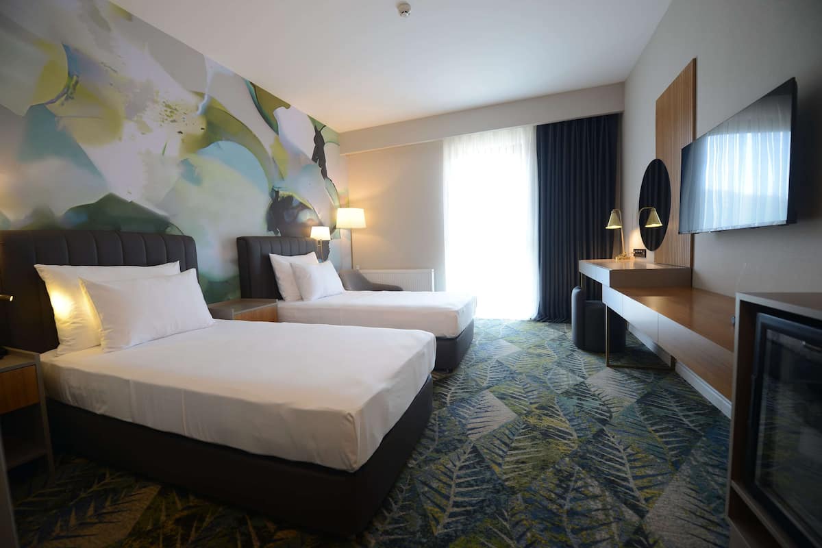 A hotel room with two beds, a large abstract painting on the wall, a flat-screen TV, and a large window with curtains.