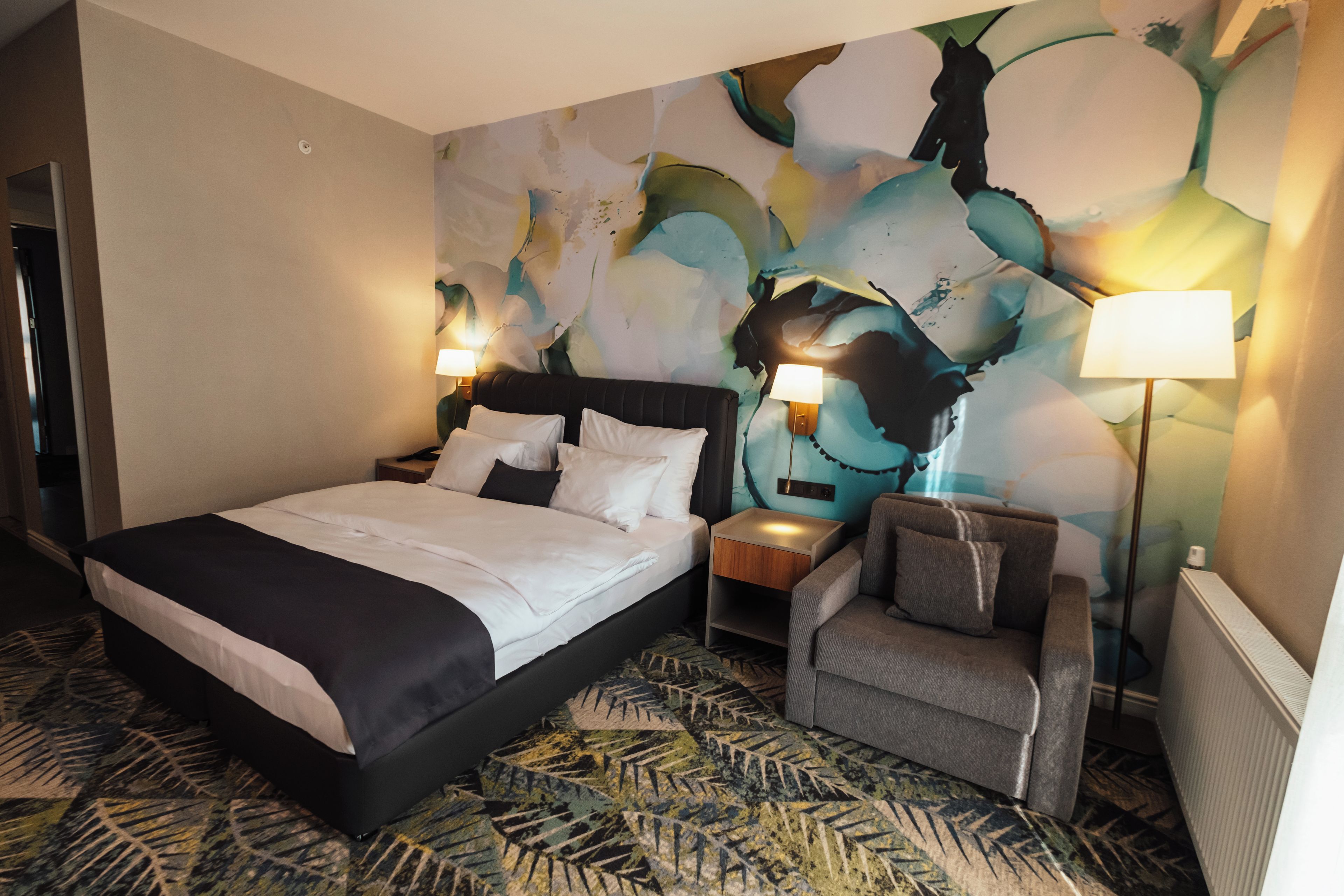 A hotel room with a large bed, two bedside tables with lamps, a chair, and a colorful abstract wall mural.