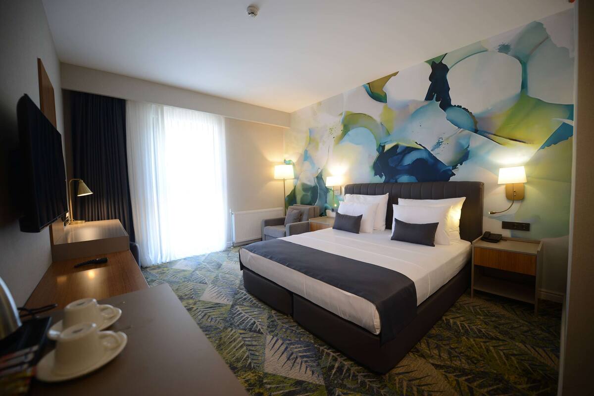 A hotel room with a large bed, two bedside tables with lamps, a desk with a chair, and a large abstract painting on the wall.