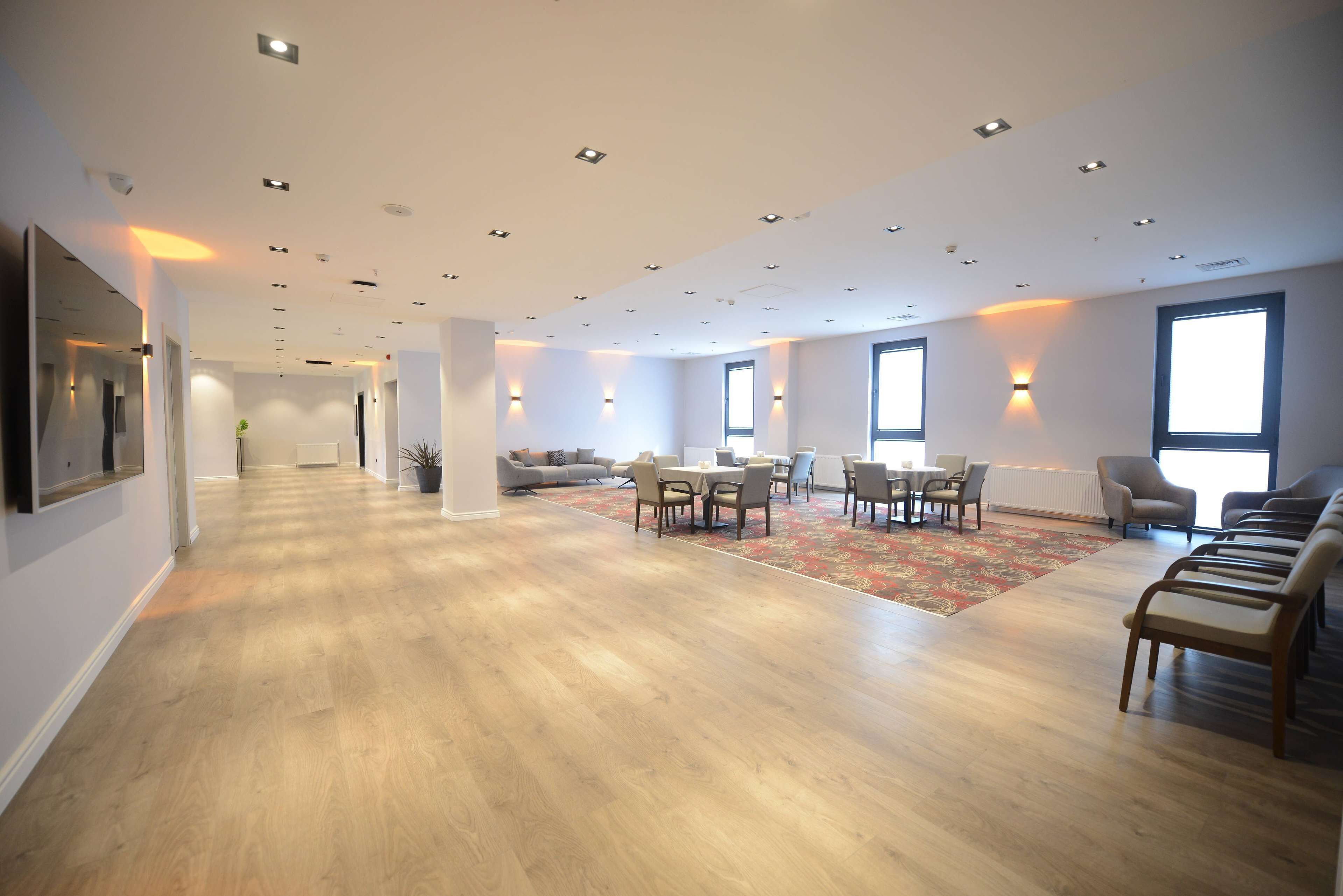 A spacious hotel lobby with a wooden floor, a seating area with tables and chairs, and large windows allowing natural light.