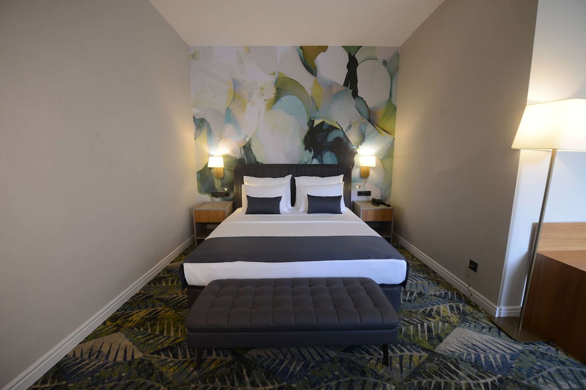 A hotel room with a large bed, two bedside lamps, a nightstand, a footboard, and a patterned carpet.
