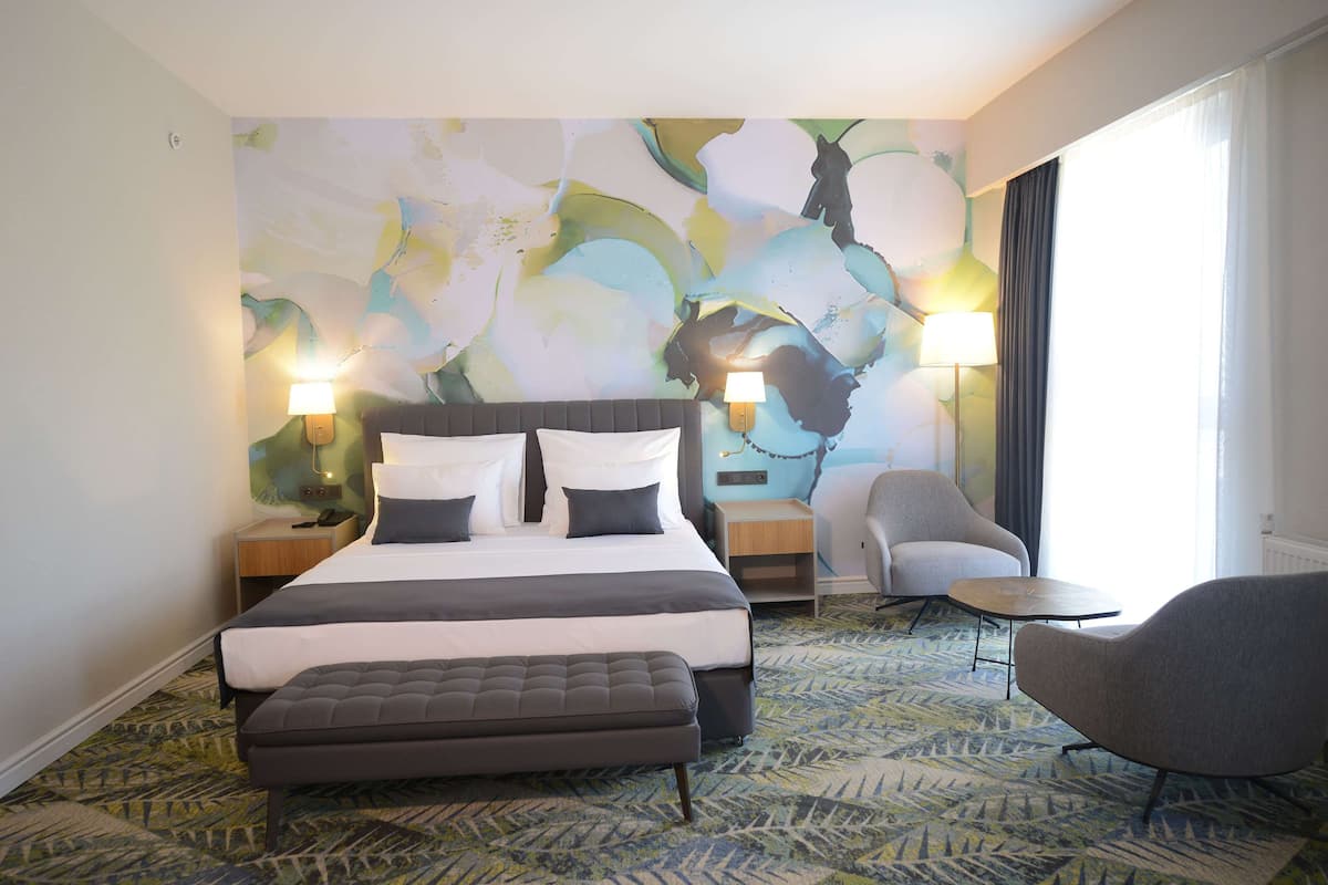 A hotel room with a large bed, two bedside tables with lamps, a grey armchair, a small round table, and a colorful abstract wall mural.