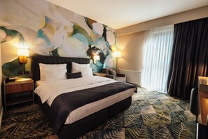 Une chambre d’hôtel avec un grand lit, deux lampes de chevet, une tête de lit, une table de chevet, une chaise, une fenêtre avec des rideaux et un mur orné d’un grand tableau abstrait.