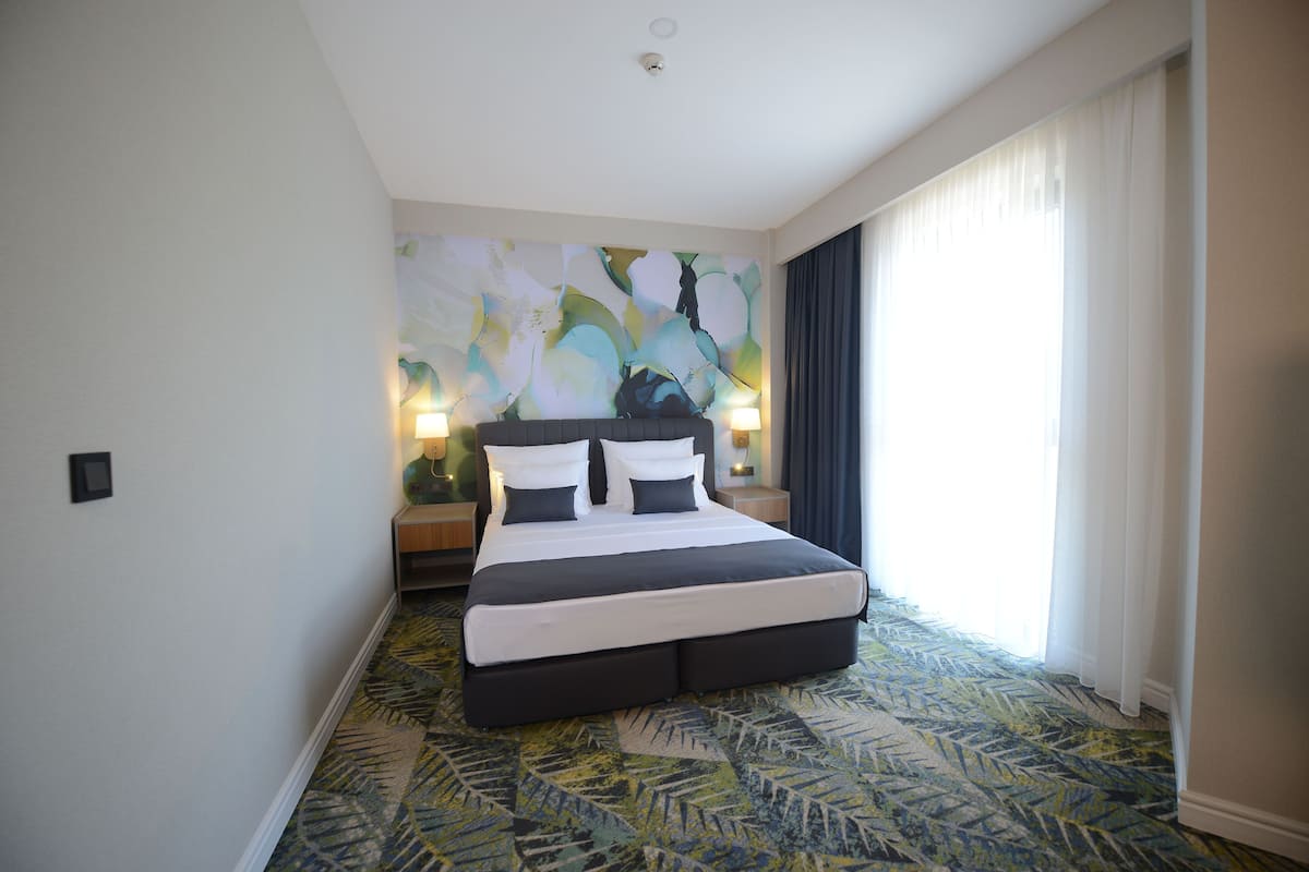 A hotel room with a large bed, two bedside lamps, a large abstract painting on the wall, and a view of the outdoors through a window with curtains.