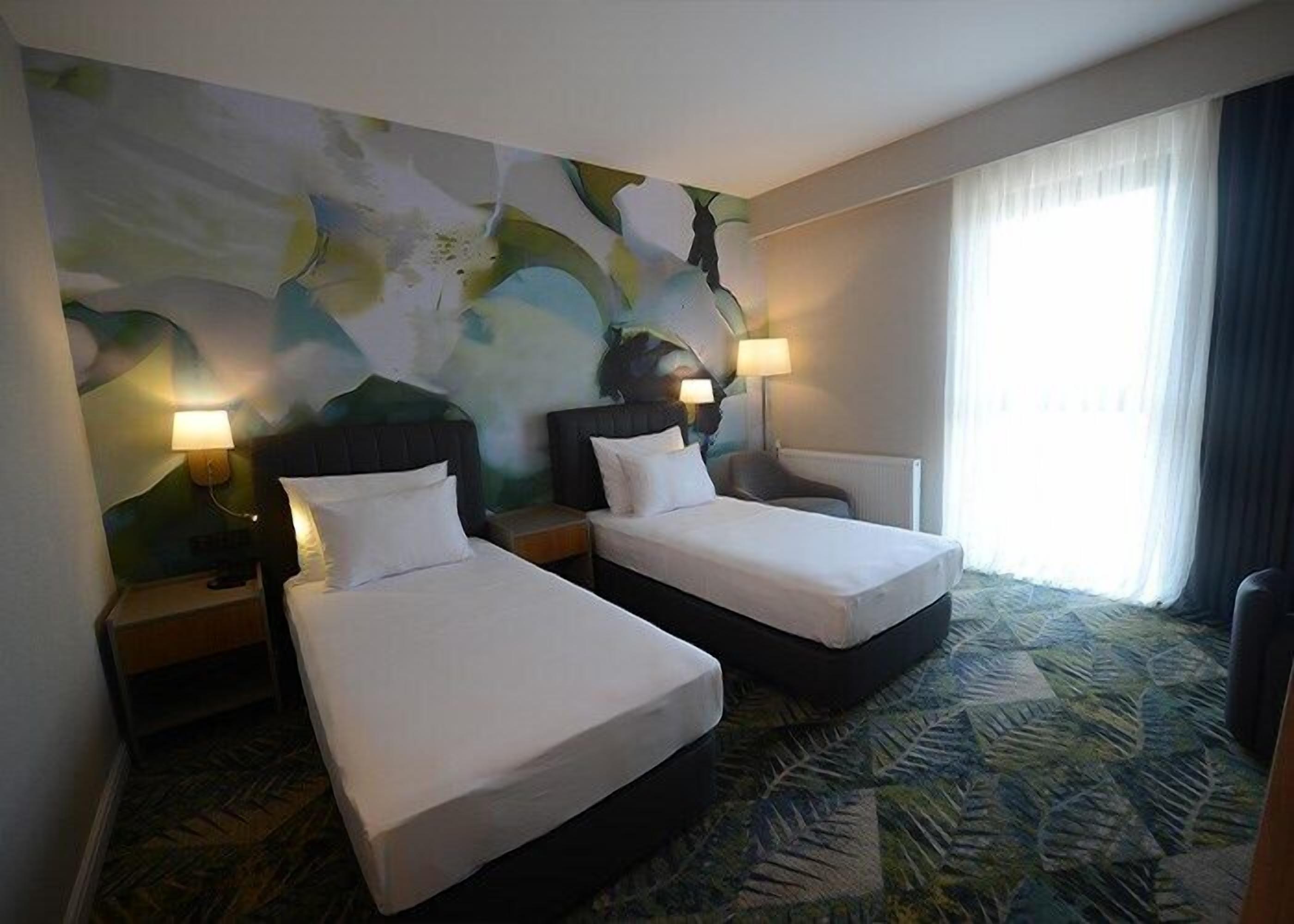 Ein Hotelzimmer mit zwei Betten, einem großen, abstrakten Wandbild und Blick ins Freie durch das Fenster.