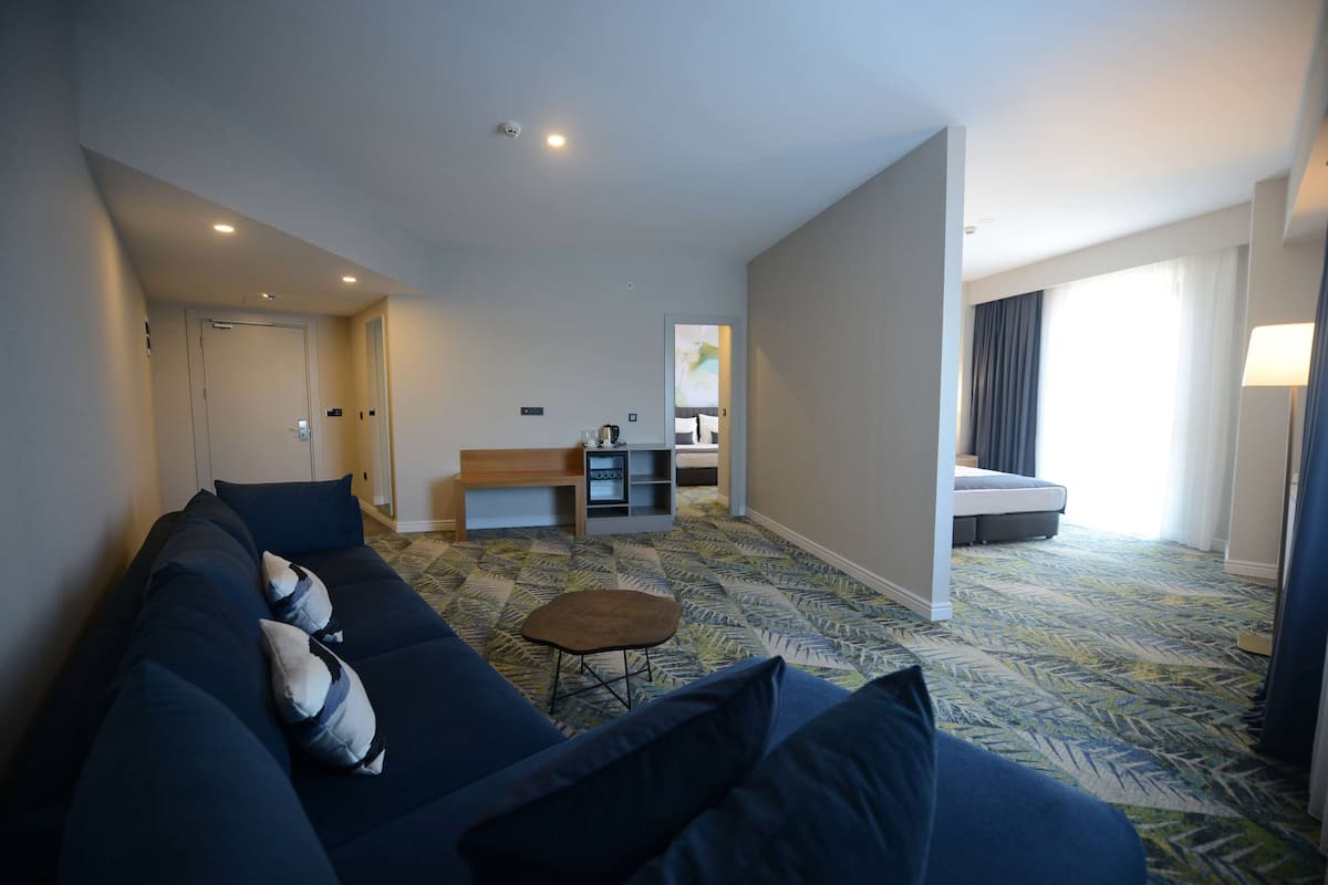 A modern hotel room with a dark blue sofa, a small wooden stool, a kitchenette, and a bed with a view of the outdoors.