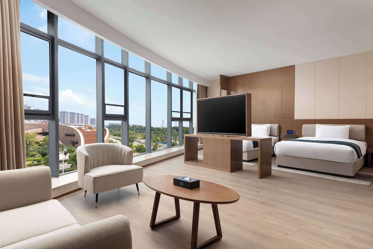 Ein modernes Hotelzimmer mit großem Fenster, Flachbildfernseher, Sofa, Bett und kleinem Tisch.