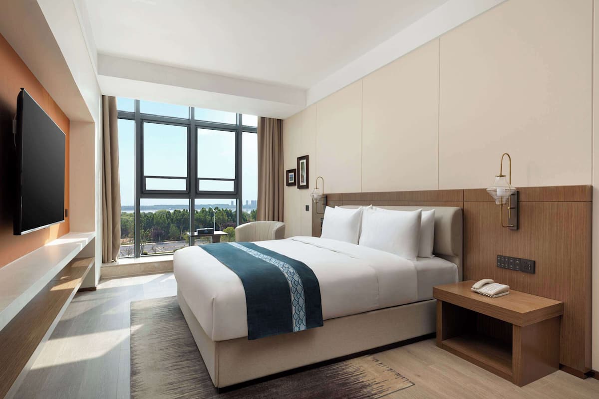 Ein modernes Hotelzimmer mit einem großen Bett, einem Nachttisch mit Telefon, einem an der Wand befestigten Fernseher und Blick ins Freie durch ein großes Fenster.