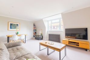 A modern living room with a sofa, a coffee table, a television, a bookshelf, and a window with a view of buildings.