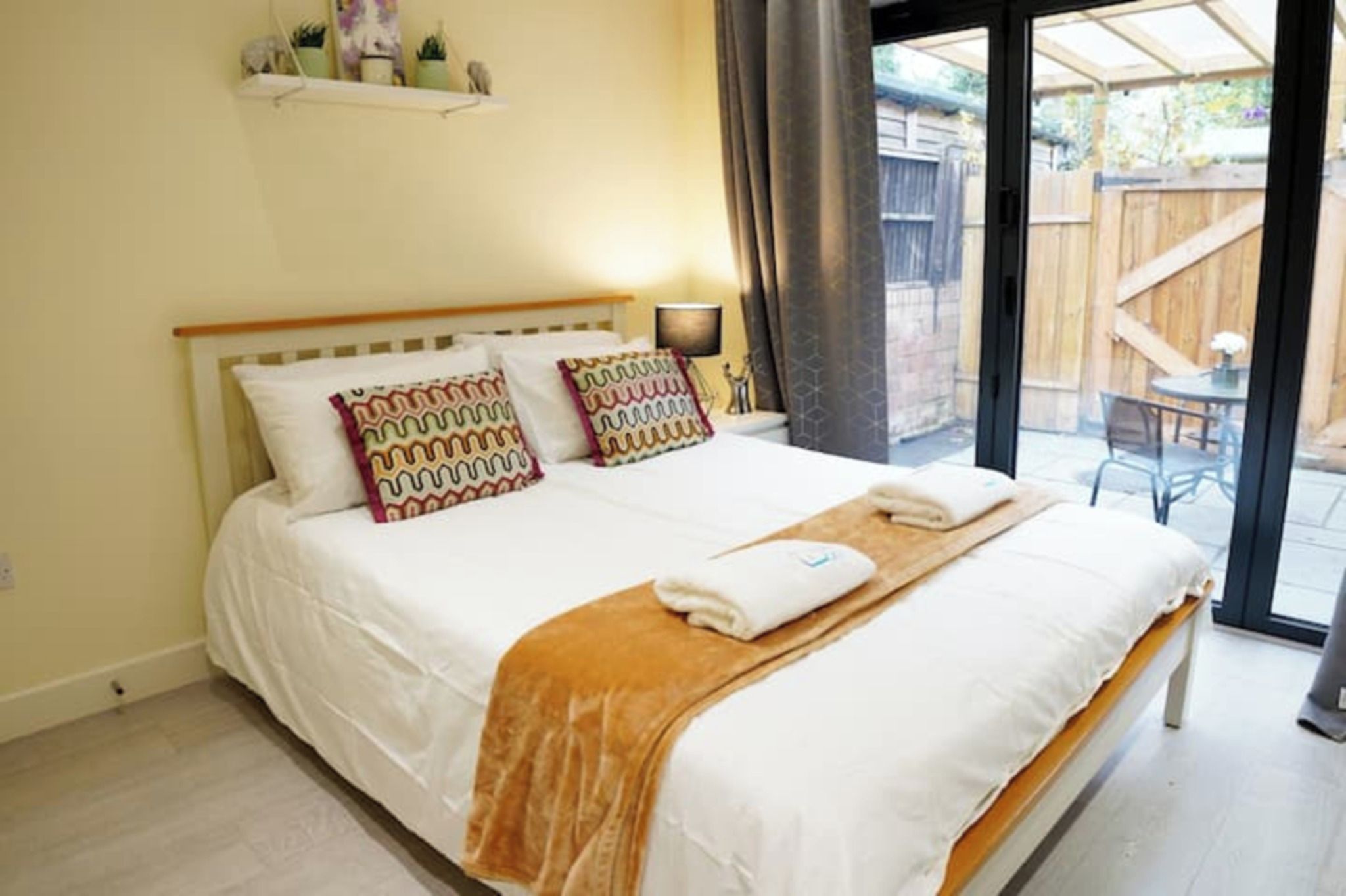 A bed with white bedding and two patterned pillows, a wooden headboard, a bedside table with a lamp, and a view of a wooden deck through a glass door.