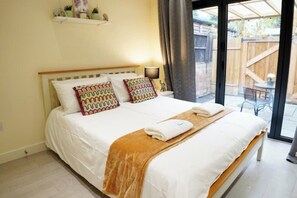 Cama com roupa de cama branca e duas almofadas estampadas, cabeceira de madeira, mesa de cabeceira com abajur e vista para um deck de madeira através de uma porta de vidro.