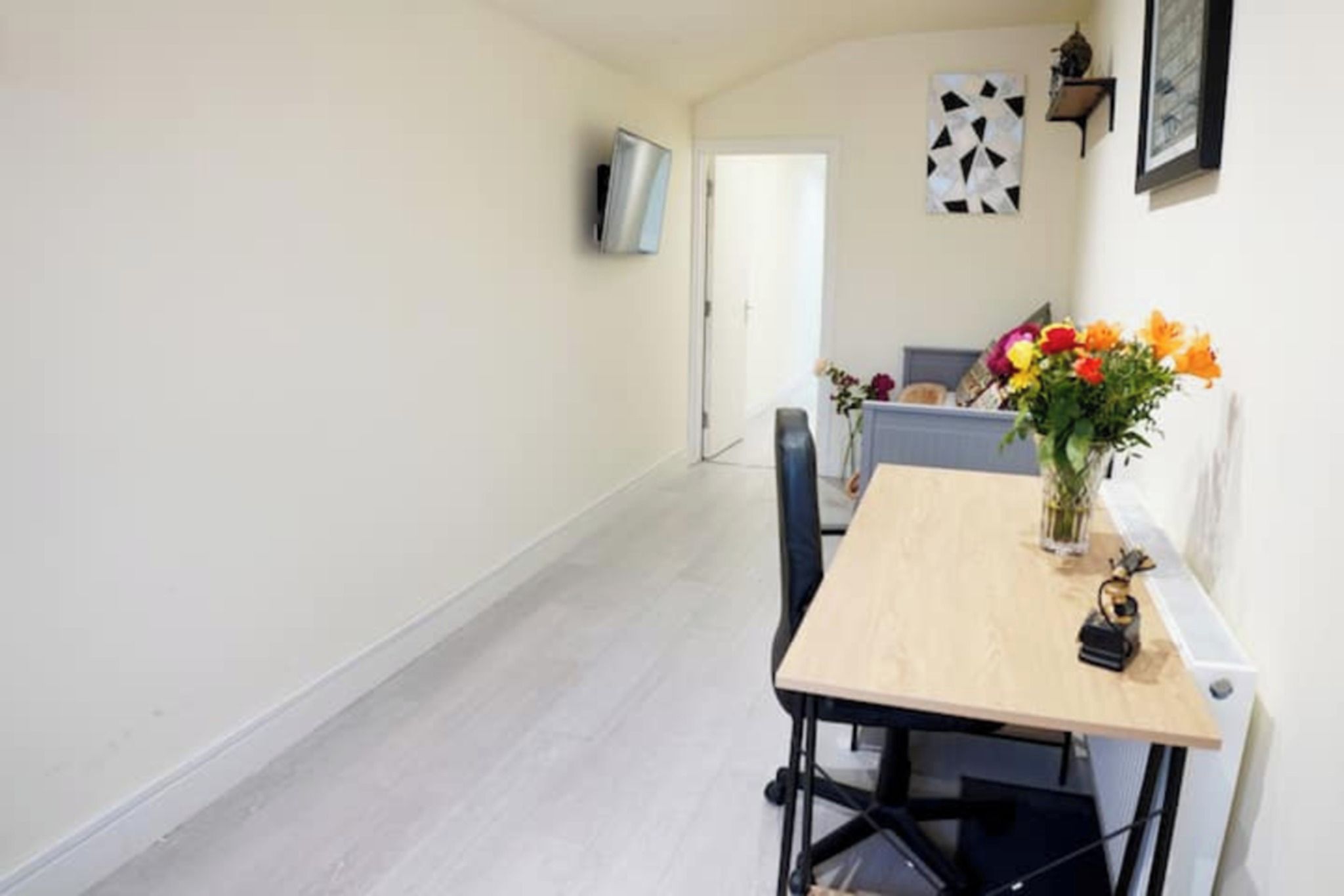 Un pasillo estrecho con una mesa de comedor y sillas, un jarrón con flores y un televisor montado en la pared.