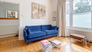 A living room with a blue sofa, a coffee table with books, a radiator, and a window with curtains. - Delightful 3BD Home with Garden in North London (London)