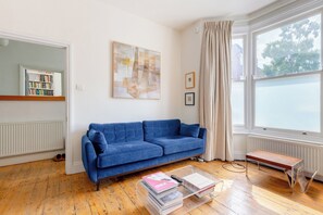 A living room with a blue sofa, a coffee table with books, a radiator, and a window with curtains. - Delightful 3BD Home with Garden in North London (London)