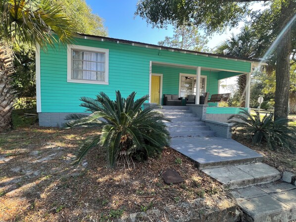 A house with a green exterior, a front porch with a bench, and a small garden with palm trees.