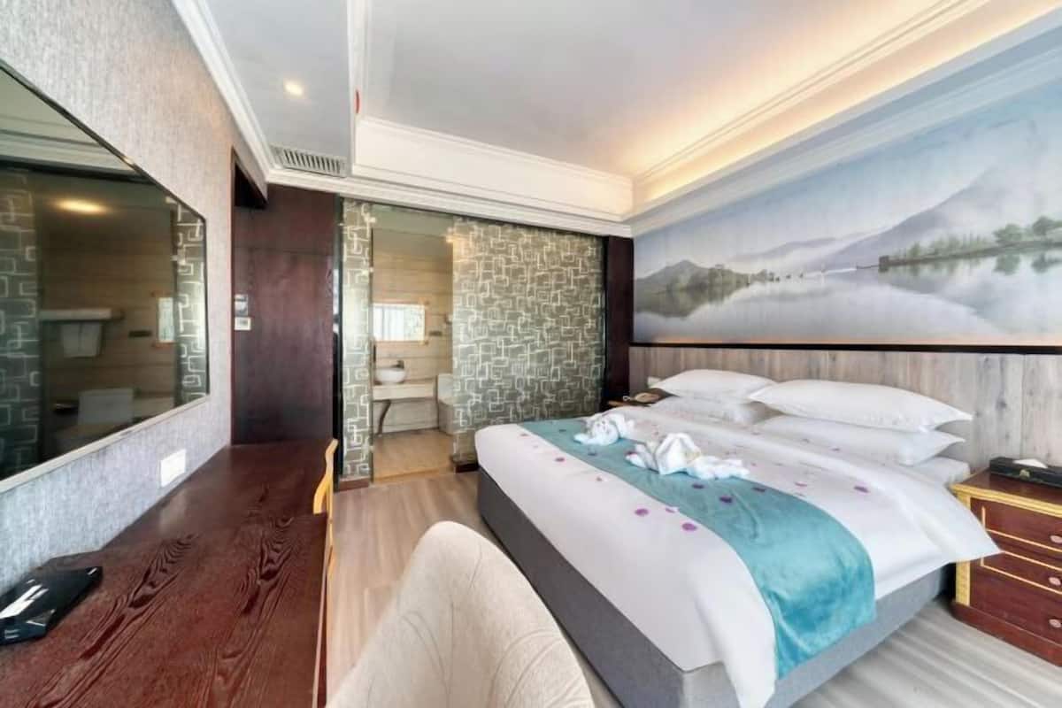 A hotel room with a large bed, a wooden desk, a stone wall, and a painting of a mountain landscape.