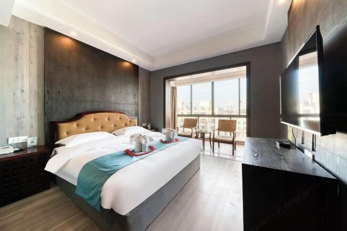 A modern hotel room with a large bed, a flat-screen TV, a city view, and a balcony with chairs.