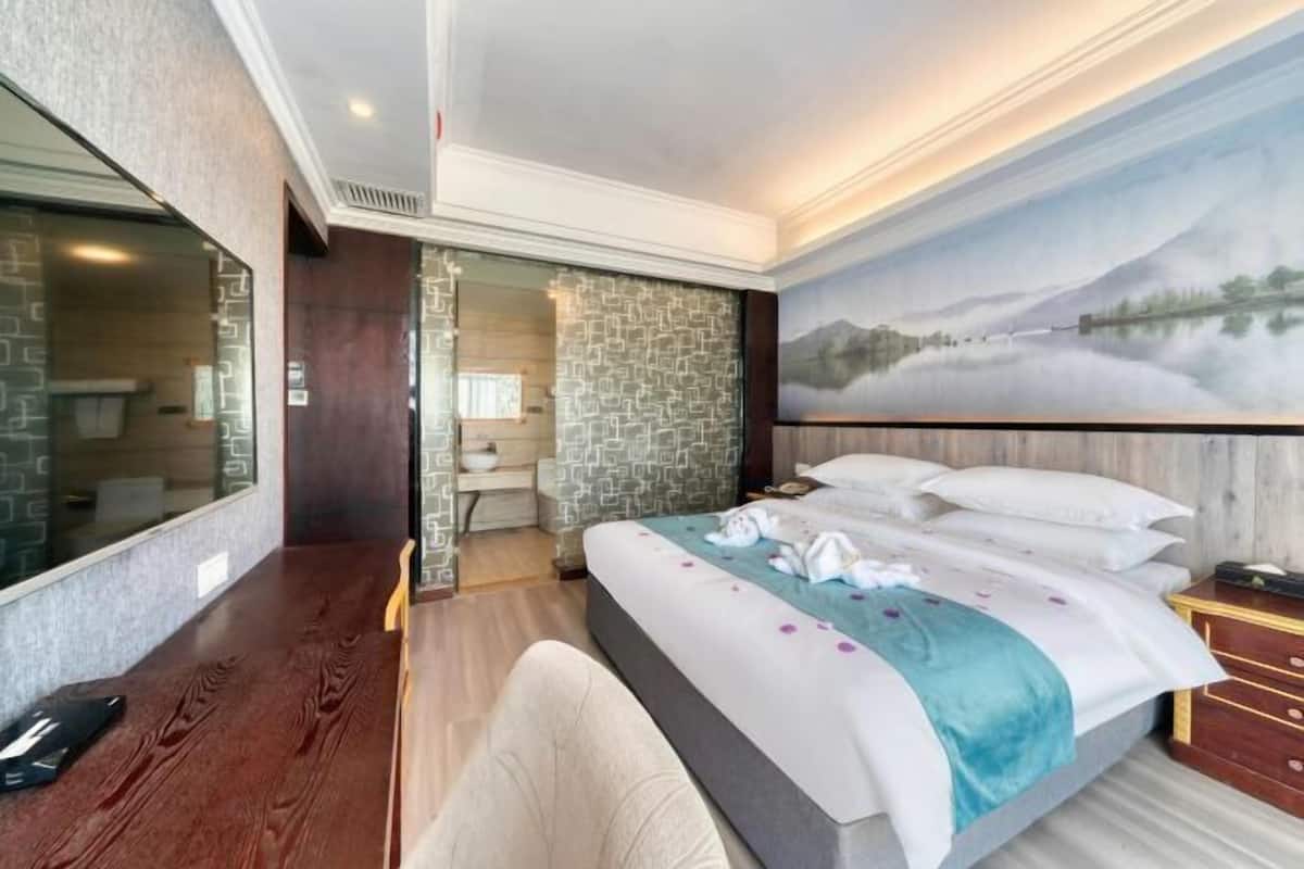A hotel room with a large bed, a wooden desk, a glass partition, and a painting of a mountain landscape.