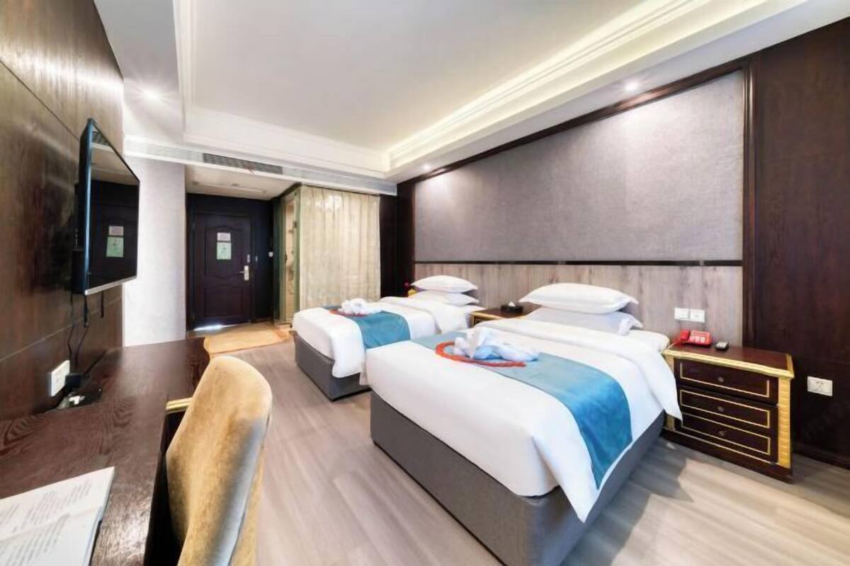 A hotel room with two beds, a desk, a chair, a TV, and a bedside table with a lamp.