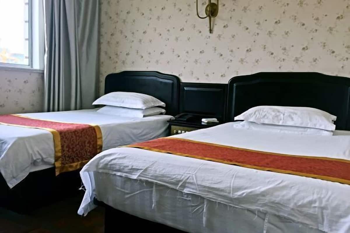 A hotel room with two single beds, each with a white bedspread and a red and gold decorative border. The room has a floral wallpaper and a window with curtains.