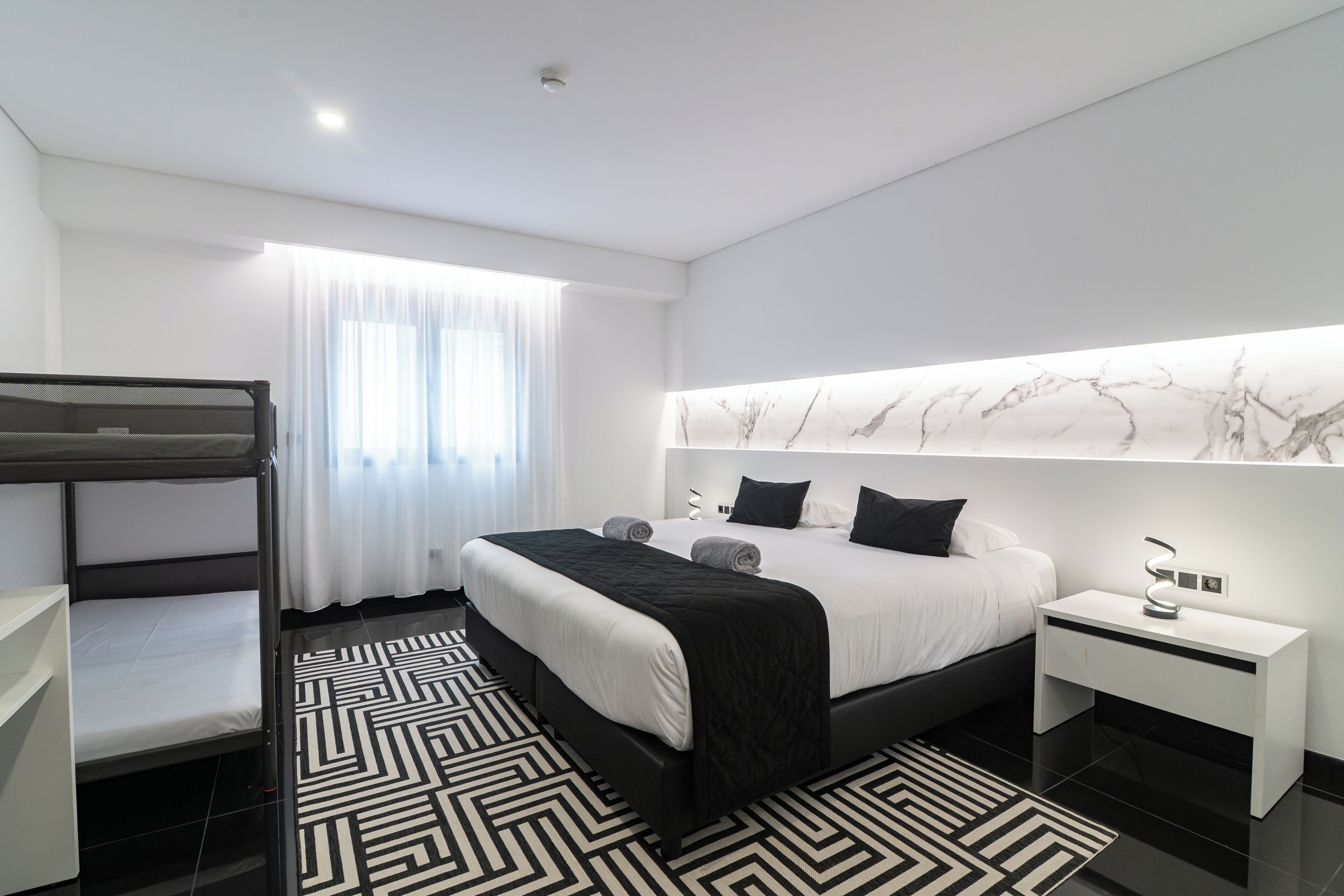 A modern hotel room with a bunk bed, a large bed with a marble headboard, a bedside table with a lamp, and a patterned rug.