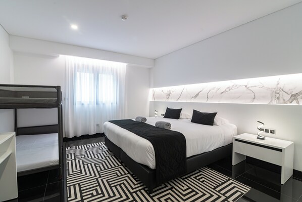 Une chambre d’hôtel moderne avec un lit superposé, un grand lit doté d’une tête de lit en marbre, une table de chevet avec une lampe et un tapis à motifs.