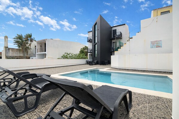 A swimming pool with lounge chairs, a modern building with balconies, and a clear sky. - The Rock Hostel (Ponta Delgada)