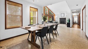 Une salle à manger spacieuse avec une longue table dressée pour quatre personnes, de grandes fenêtres équipées de stores en bois, une cheminée et un couloir menant à d’autres pièces.