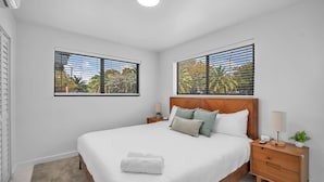 Un dormitorio moderno con cabecera de madera, una cama grande con ropa de cama blanca, dos mesitas de noche con lámparas y vistas a palmeras a través de las ventanas.