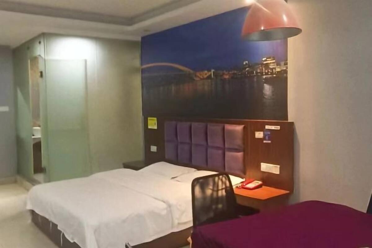 Habitación de hotel con cama, escritorio, silla, mesita de noche, lámpara y vistas a la ciudad.