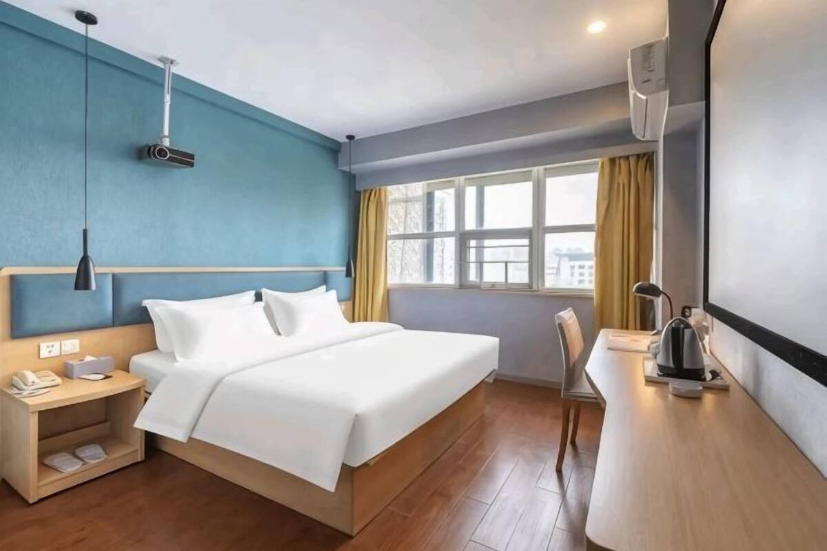 Una habitación de hotel con cama, mesita de noche, lámpara y ventana con cortinas.