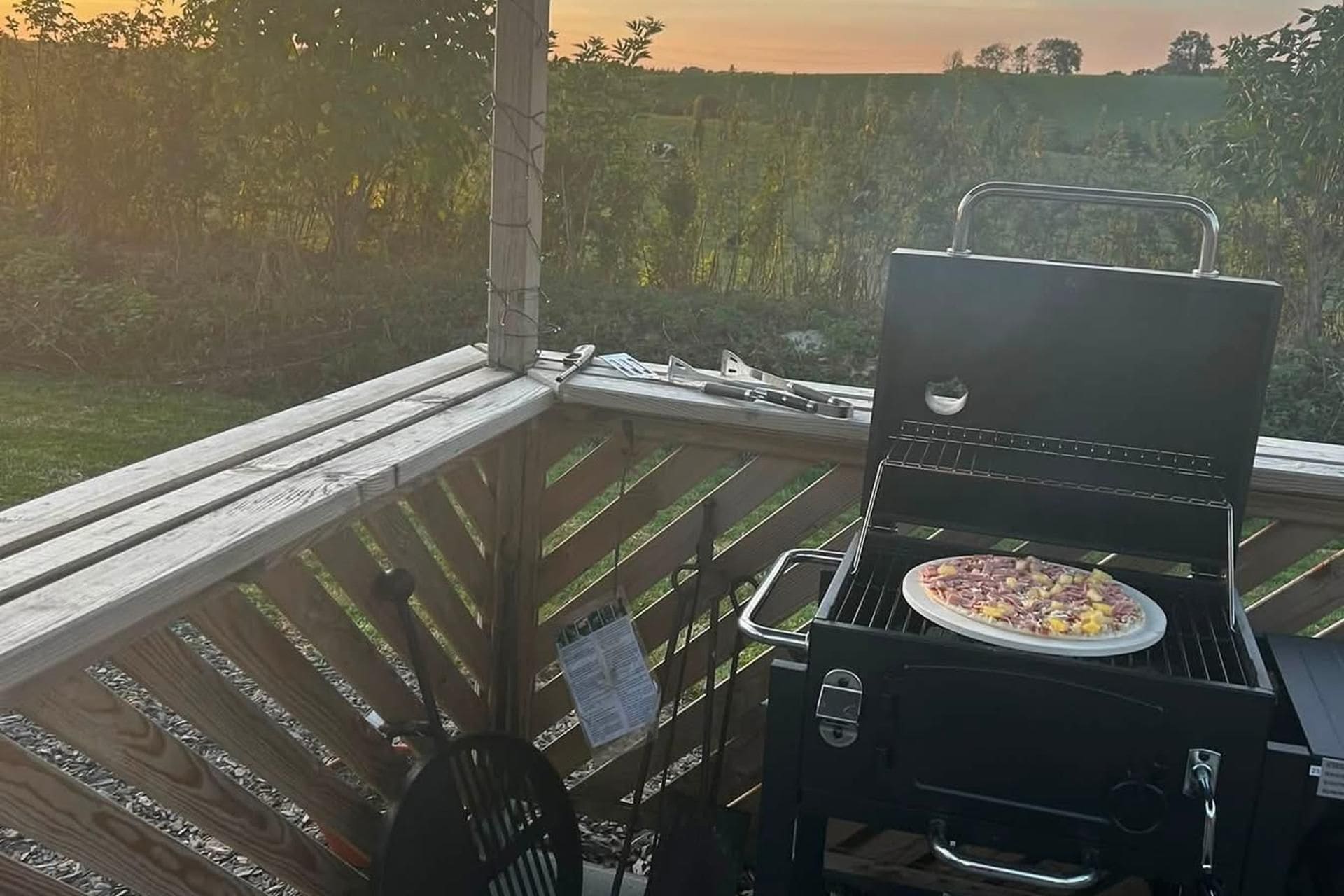 A barbecue grill with a pizza on it, located on a wooden deck with a view of a field.