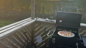 A barbecue grill with a pizza on it, located on a wooden deck with a view of a field.