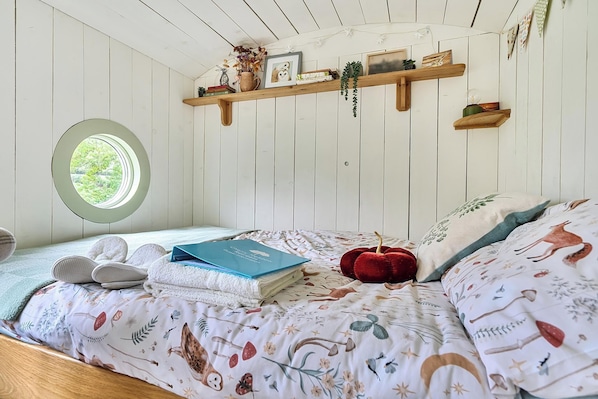 A bedroom with a bed, a round window, wooden shelves, and a view of greenery outside.