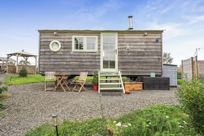 A wooden cabin with a front door, windows, and a chimney. There is a small porch with a table and chairs, and a ladder leading to the door. - Cosy Luxury Owls Roost Hut With Private Hot Tub (Umberleigh)