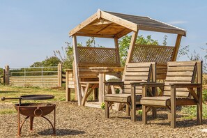 Un mirador de madera con techo de celosía, rodeado de sillas de madera y un hogar a fuego de metal.