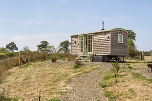 Una pequeña casa de madera con porche y chimenea, rodeada por un camino de grava y vegetación.