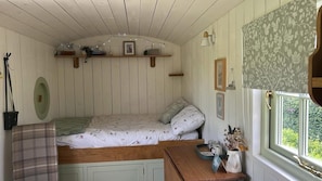 A cozy bedroom with a bed, a small table, a window with a view, and a shelf with decorative items. - Cosy Luxury Foxglove Hut With Private Fire Bowl (Umberleigh)