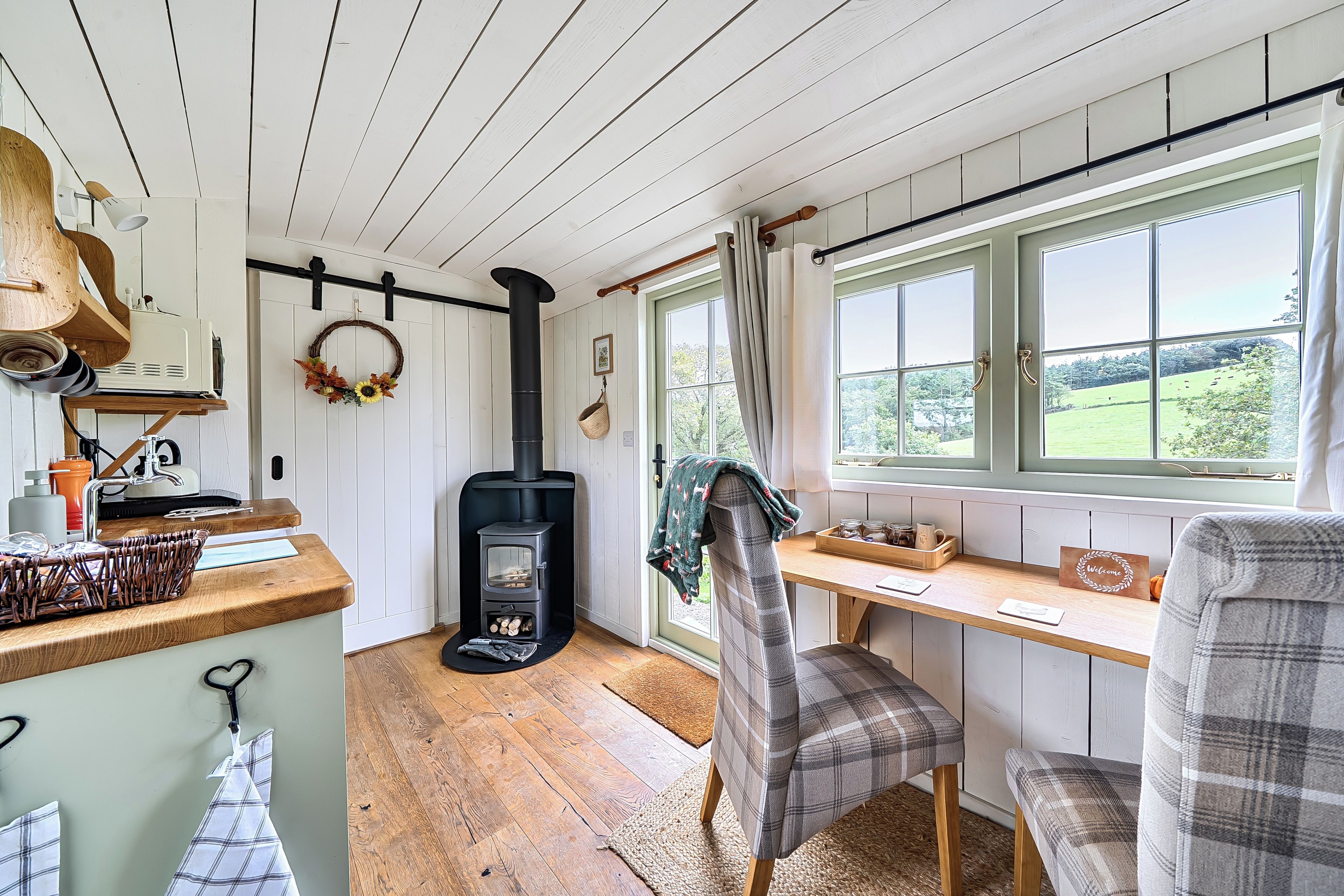 A cozy room with a wood-burning stove, a dining table with chairs, a window with a view of the countryside, and a kitchen area with a hanging wreath.