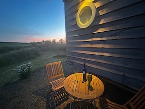 Et udendørs siddeområde med et bord, stole, en flaske og et glas foran en trævæg med et vindue.