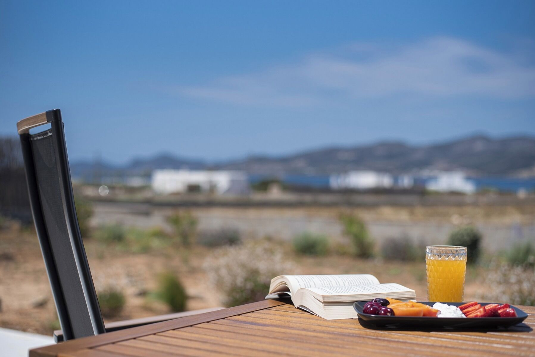 A table with a book, fruit, and a glass of orange juice, overlooking a landscape with buildings.